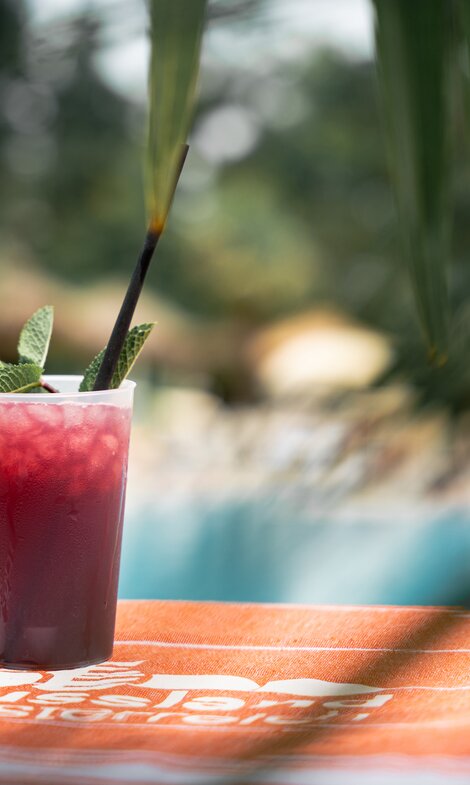 Roter Cocktail auf orangem Strandtuch, bedeckt von Palmen und im Hintergrund ein Pool | © Spa Resort Geinberg / Tobi Gast