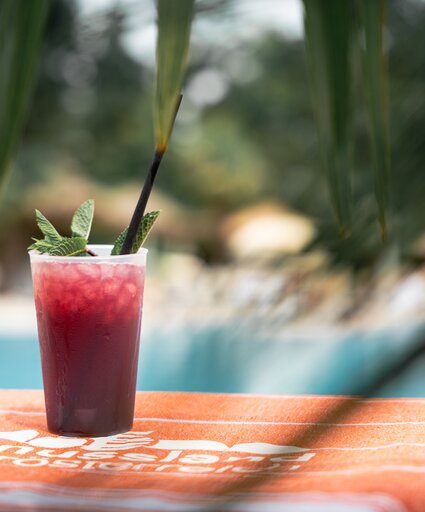 Roter Cocktail auf orangem Strandtuch, bedeckt von Palmen und im Hintergrund ein Pool | © Spa Resort Geinberg / Tobi Gast