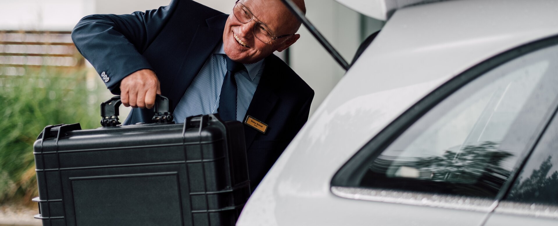 Älterer Mann im Anzug lächelt und hievt einen Koffer aus dem Kofferraum eines Autos | © Spa Resort Geinberg / Chris Perkles