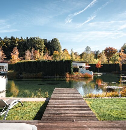 Herbstliche Außenansicht der Geinberg5 Private Spa Villas mit See, Steg und bunten Bäumen unter blauem Himmel | © Spa Resort Geinberg / Tobi Gast