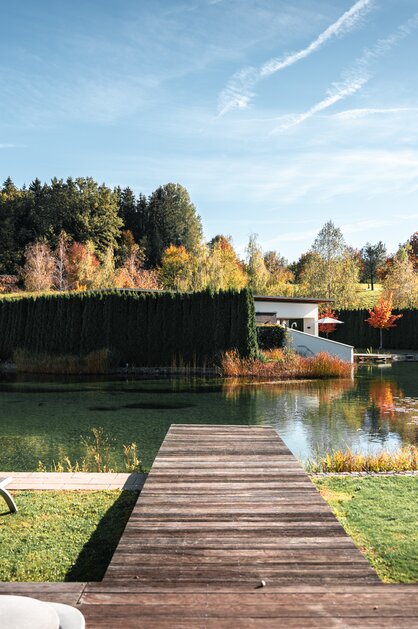 Herbstliche Außenansicht der Geinberg5 Private Spa Villas mit See, Steg und bunten Bäumen unter blauem Himmel | © Spa Resort Geinberg / Tobi Gast