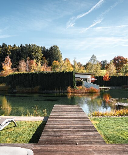 Herbstliche Außenansicht der Geinberg5 Private Spa Villas mit See, Steg und bunten Bäumen unter blauem Himmel | © Spa Resort Geinberg / Tobi Gast