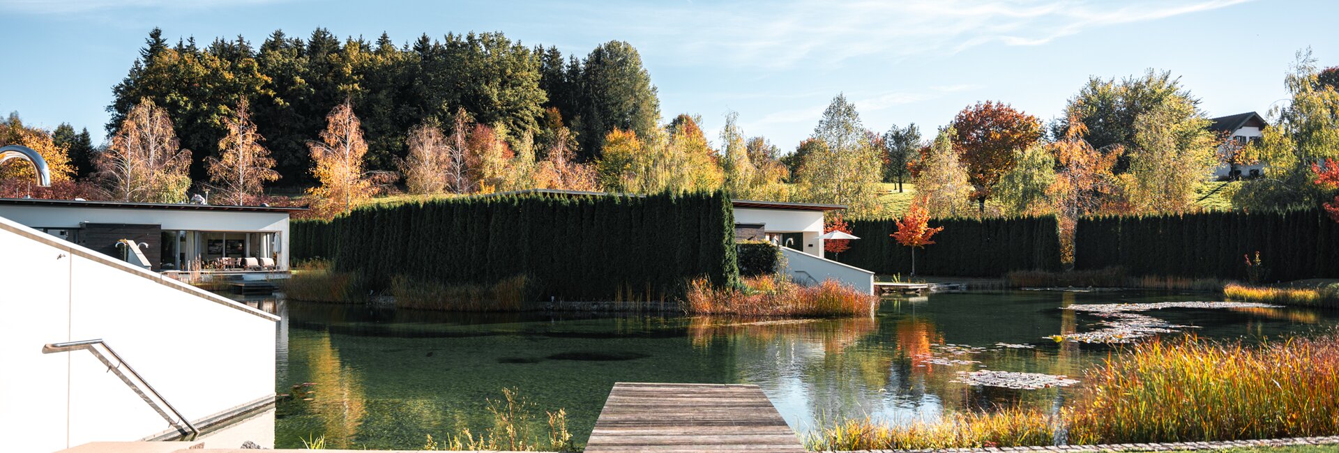 Herbstliche Außenansicht der Geinberg5 Private Spa Villas mit See, Steg und bunten Bäumen unter blauem Himmel | © Spa Resort Geinberg / Tobi Gast