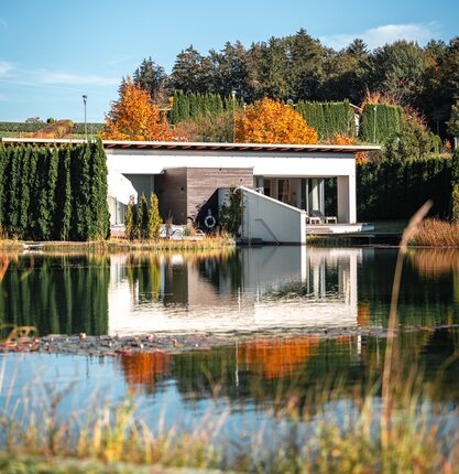 © Spa Resort Geinberg / Tobi Gast Moderne Villa am See, umgeben von herbstlicher Natur und Spiegelungen im Wasser | © Spa Resort Geinberg / Tobi Gast