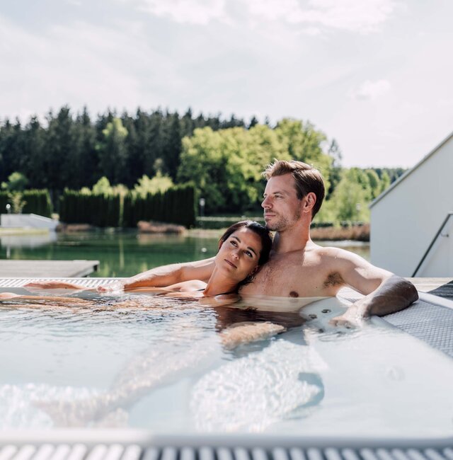 Paar entspannt im Whirlpool auf einer Terrasse mit Blick auf See und Wald