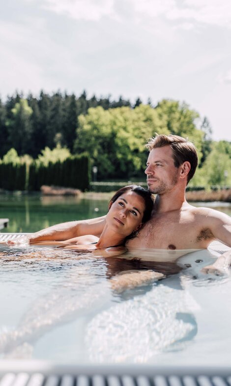 Paar entspannt im Whirlpool auf einer Terrasse mit Blick auf See und Wald