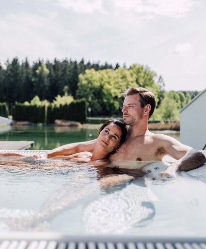 Paar entspannt im Whirlpool auf einer Terrasse mit Blick auf See und Wald