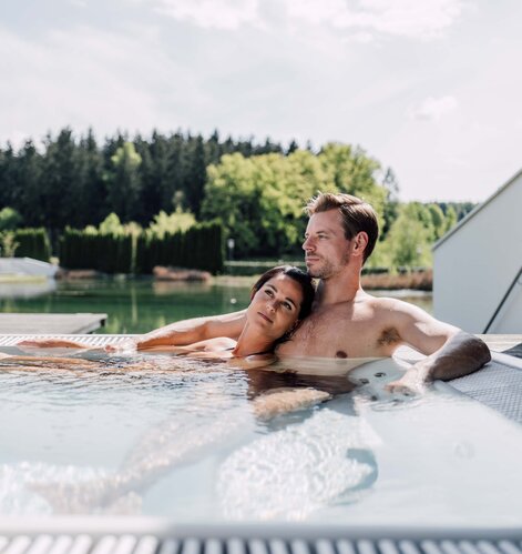 Paar entspannt im Whirlpool auf einer Terrasse mit Blick auf See und Wald