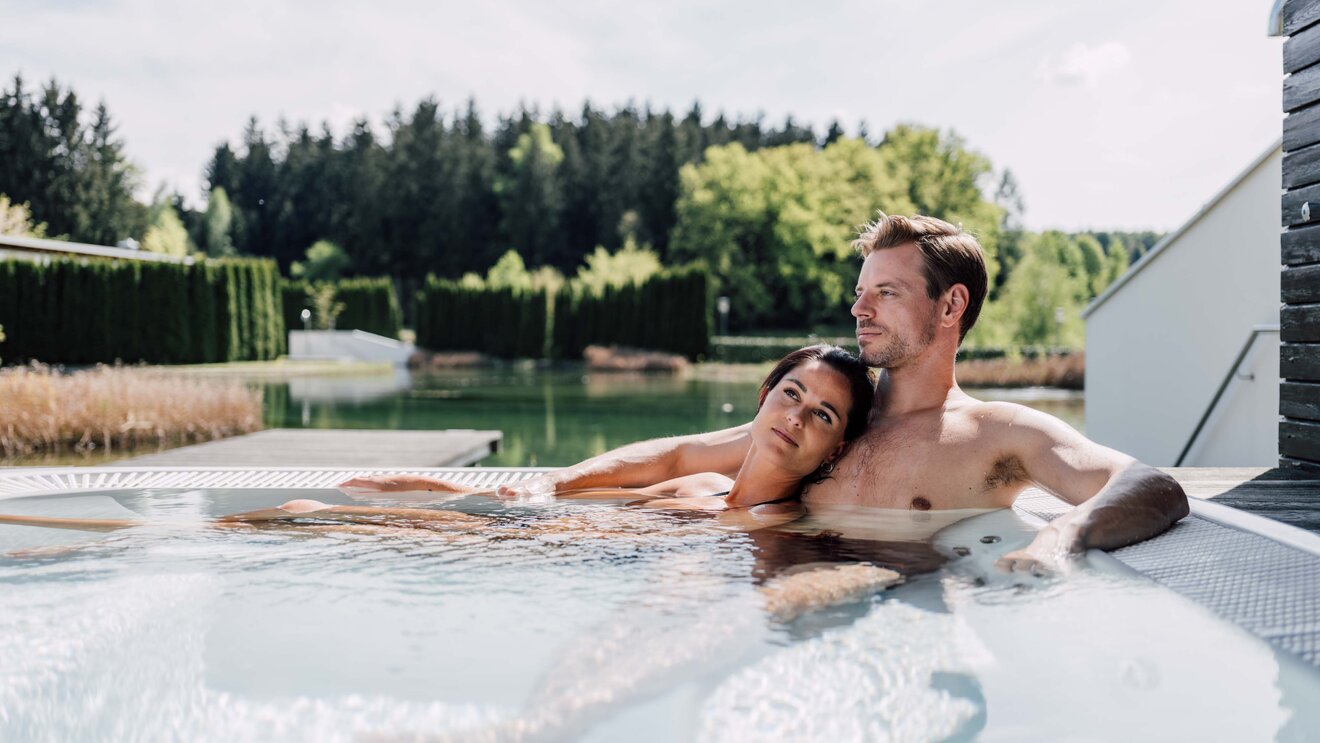 Paar entspannt im Whirlpool auf einer Terrasse mit Blick auf See und Wald