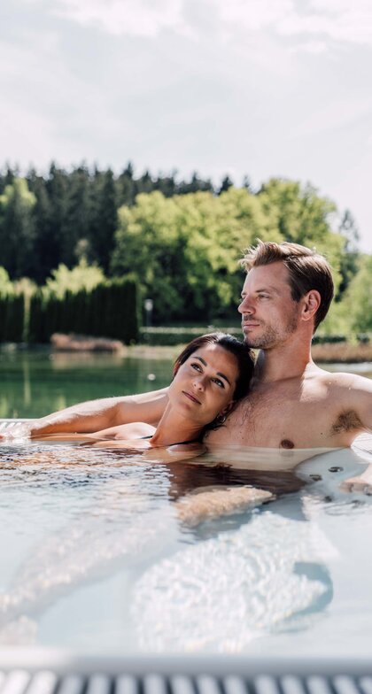 Paar entspannt im Whirlpool auf einer Terrasse mit Blick auf See und Wald