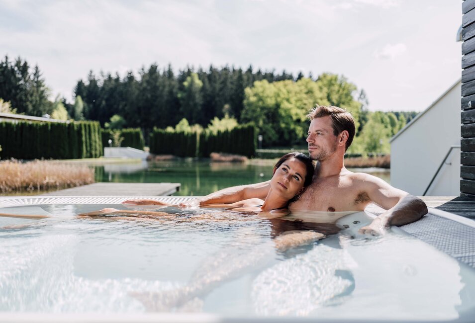 Paar entspannt im Whirlpool auf einer Terrasse mit Blick auf See und Wald