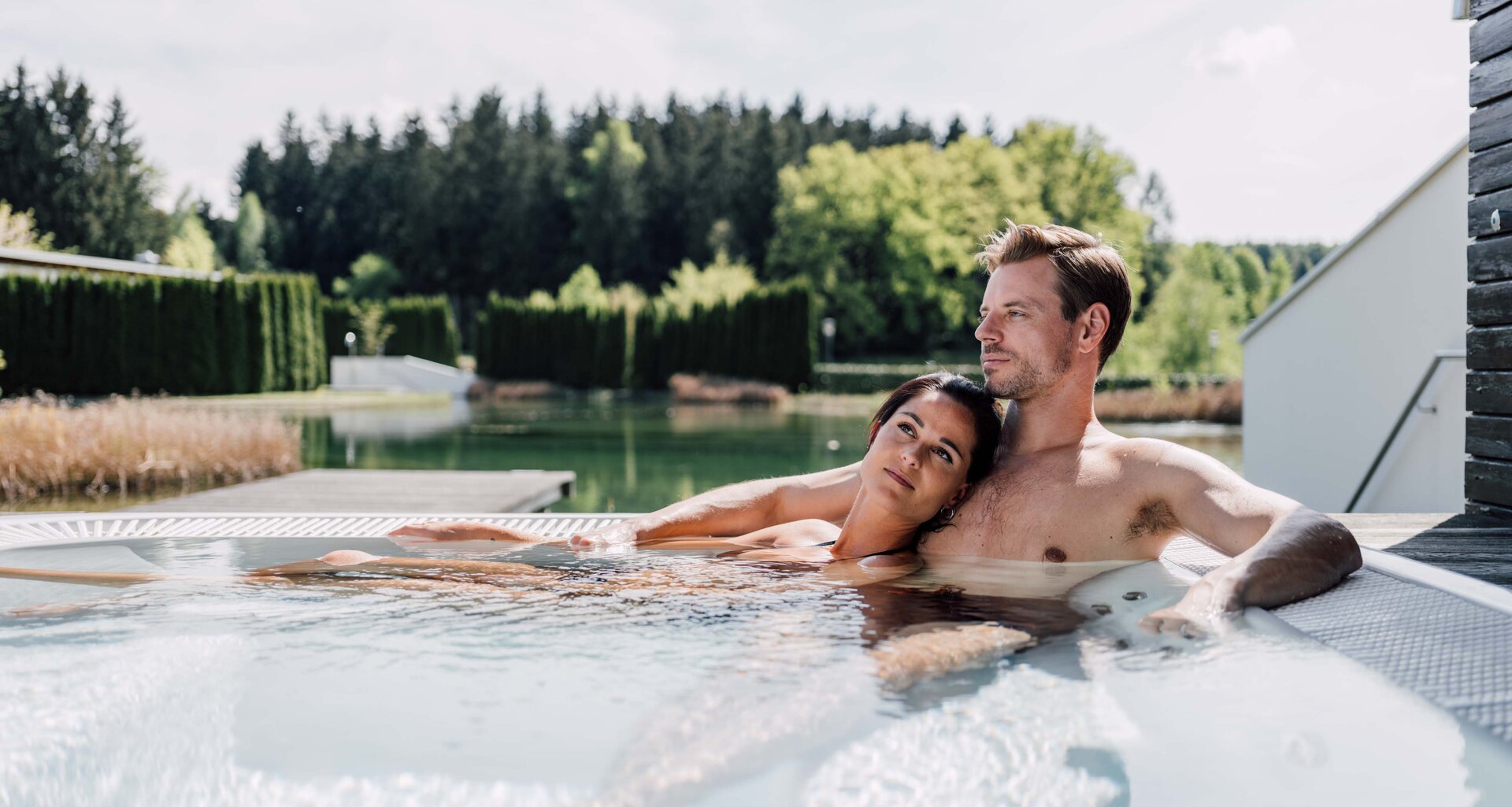 Paar entspannt im Whirlpool auf einer Terrasse mit Blick auf See und Wald