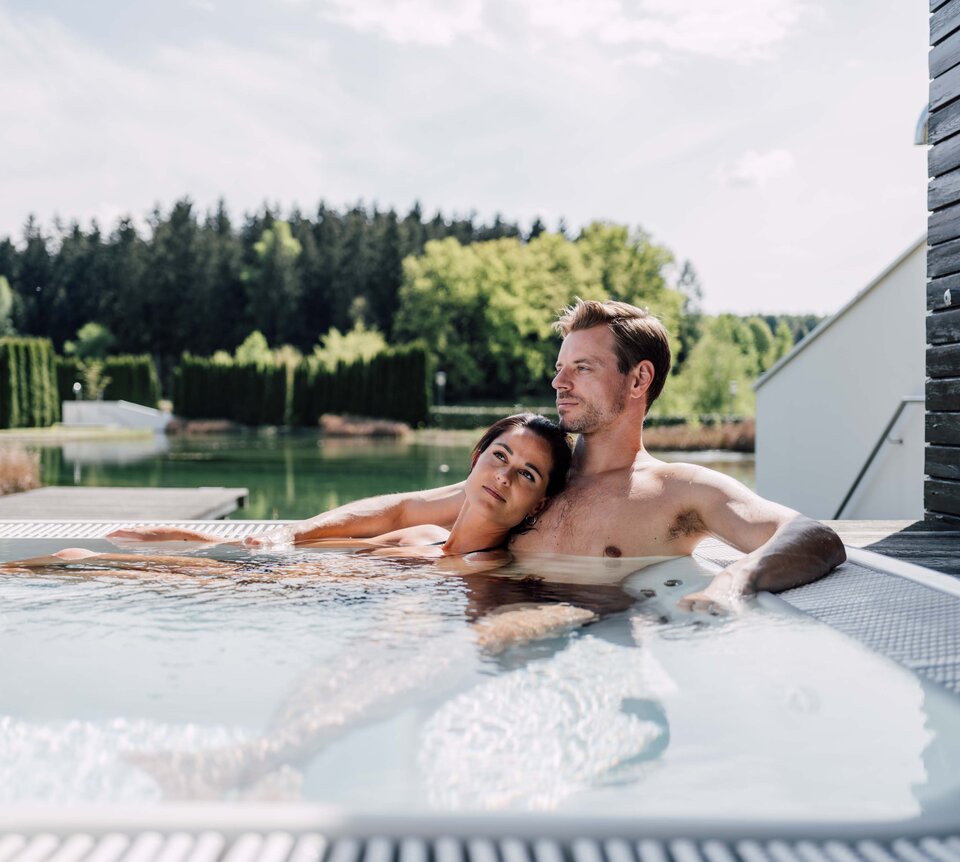 Paar entspannt im Whirlpool auf einer Terrasse mit Blick auf See und Wald