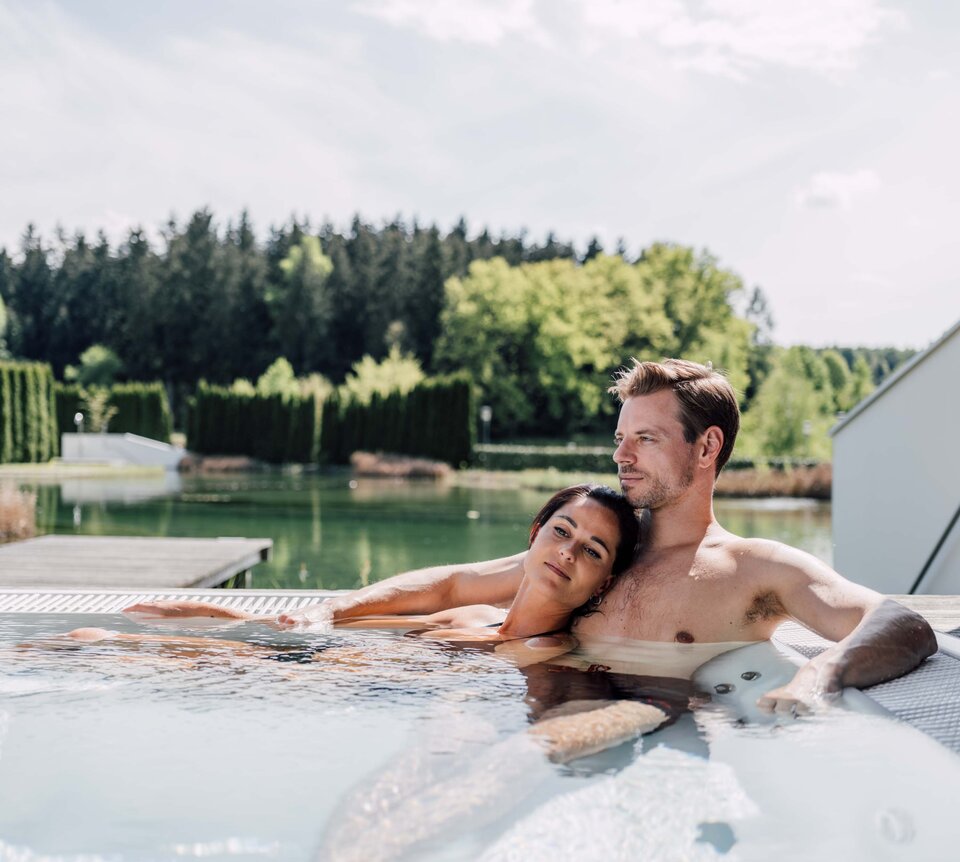 Paar entspannt im Whirlpool einer Terrasse mit Blick auf einen See und grüne Landschaft