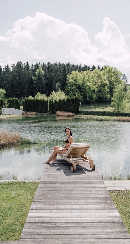 Frau im Bikini sitzt auf Liege am Badesteg vor See und Wald | © Spa Resort Geinberg / Chris Perkles