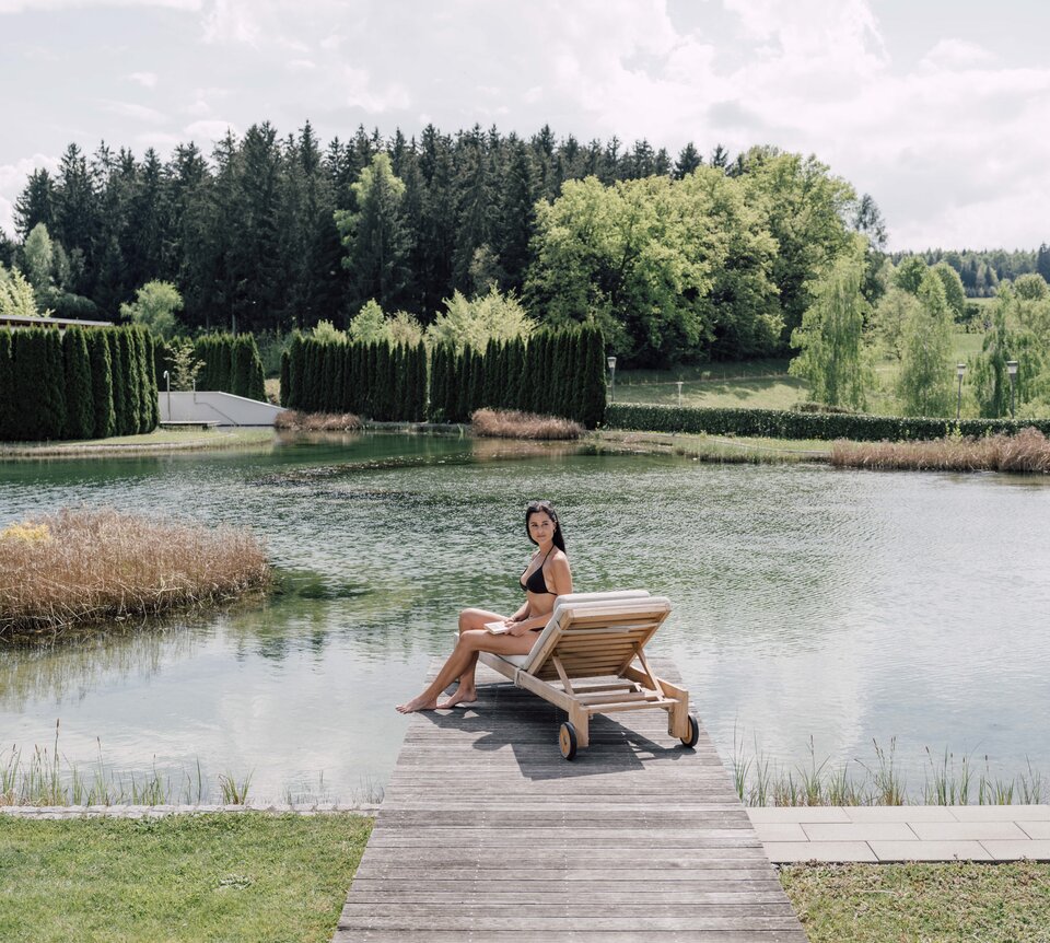 Frau im Bikini sitzt auf Liege am Badesteg vor See und Wald | © Spa Resort Geinberg / Chris Perkles