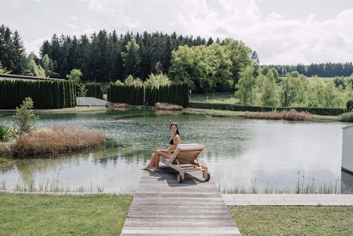 Frau auf Liege am Badesteg eines Sees vor Villen und Wald | © Spa Resort Geinberg / Chris Perkles