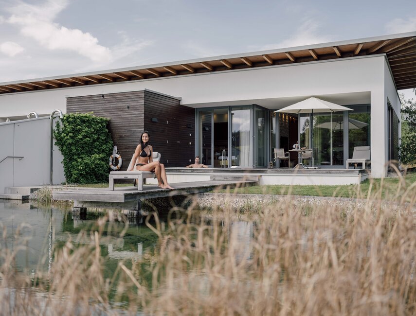 Frau im Bikini sitzt auf Badesteg vor moderner Villa am Wasser | © Spa Resort Geinberg / Chris Perkles