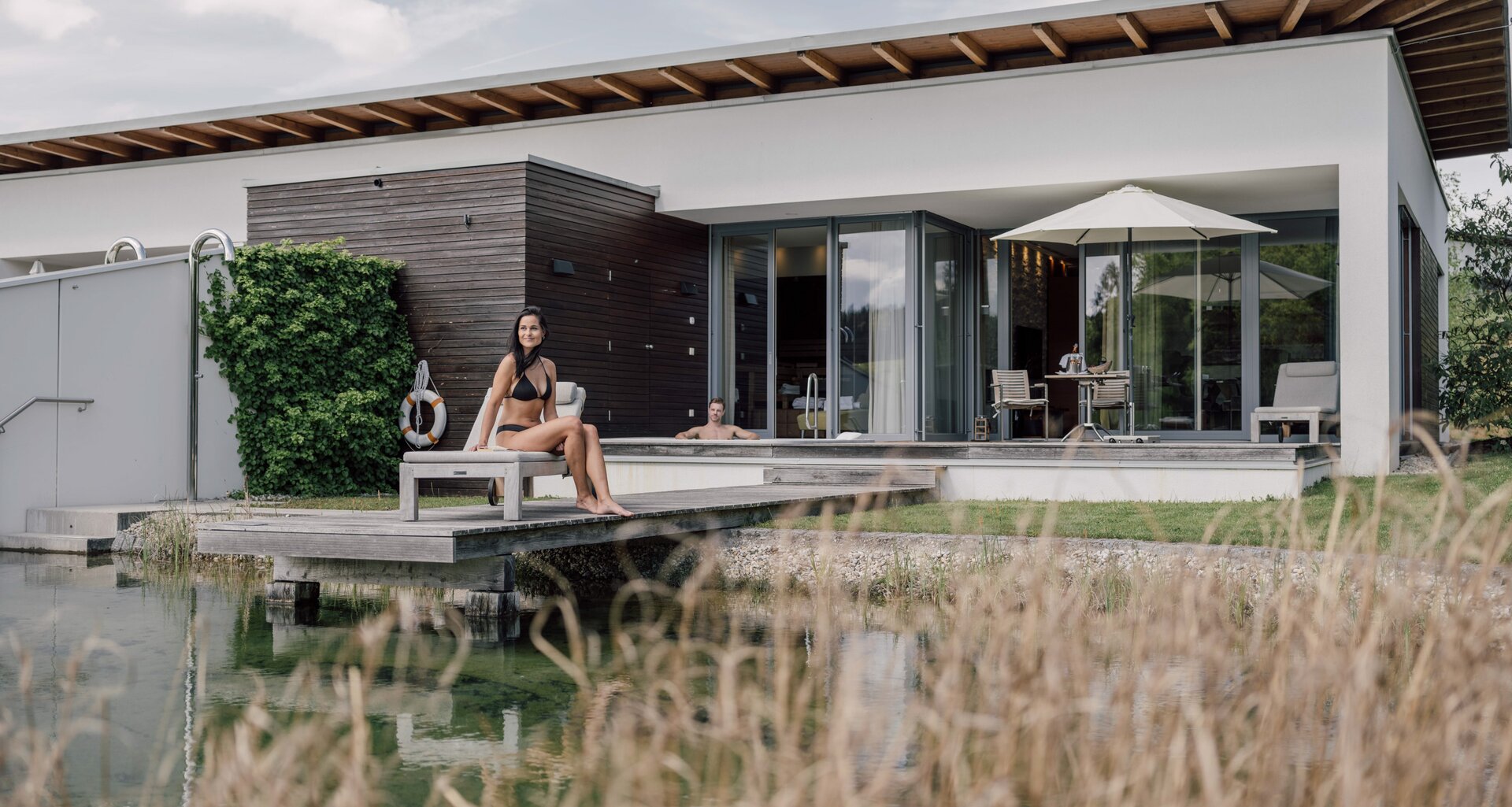 © Spa Resort Geinberg / Chris Perkles Frau im Bikini sitzt auf Badesteg vor moderner Villa am Wasser | © Spa Resort Geinberg / Chris Perkles