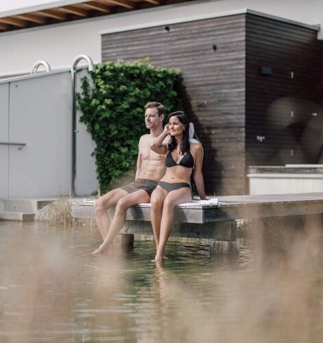 Paar sitzt barfuß auf Holzsteg am Wasser vor Gebäude mit Grünbewuchs | © Spa Resort Geinberg / Chris Perkles