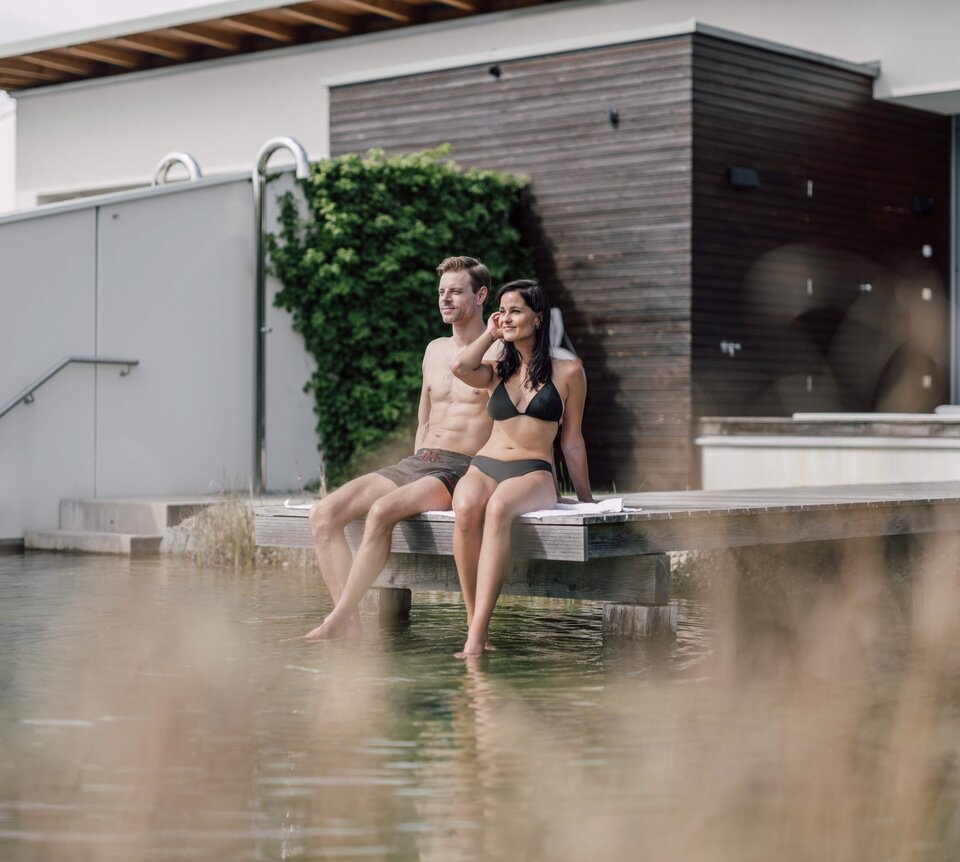 Paar sitzt barfuß auf Holzsteg am Wasser vor Gebäude mit Grünbewuchs | © Spa Resort Geinberg / Chris Perkles