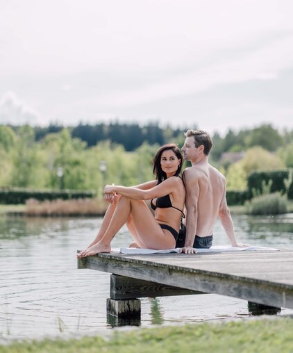 Paar sitzt Rücken an Rücken auf Badesteg am See | © Spa Resort Geinberg / Chris Perkles