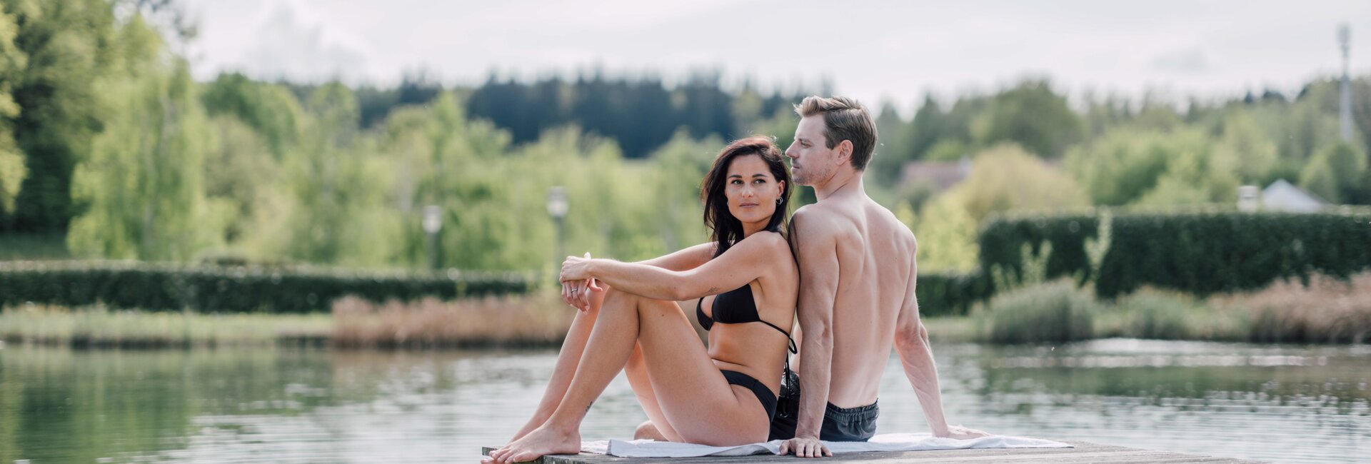 Paar sitzt Rücken an Rücken auf Badesteg am See | © Spa Resort Geinberg / Chris Perkles