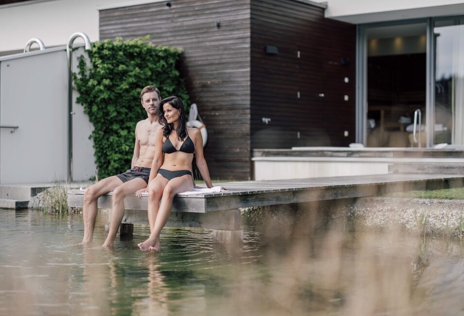 Paar sitzt barfuß auf Holzsteg am Wasser vor Spa-Villa | © Spa Resort Geinberg / Chris Perkles