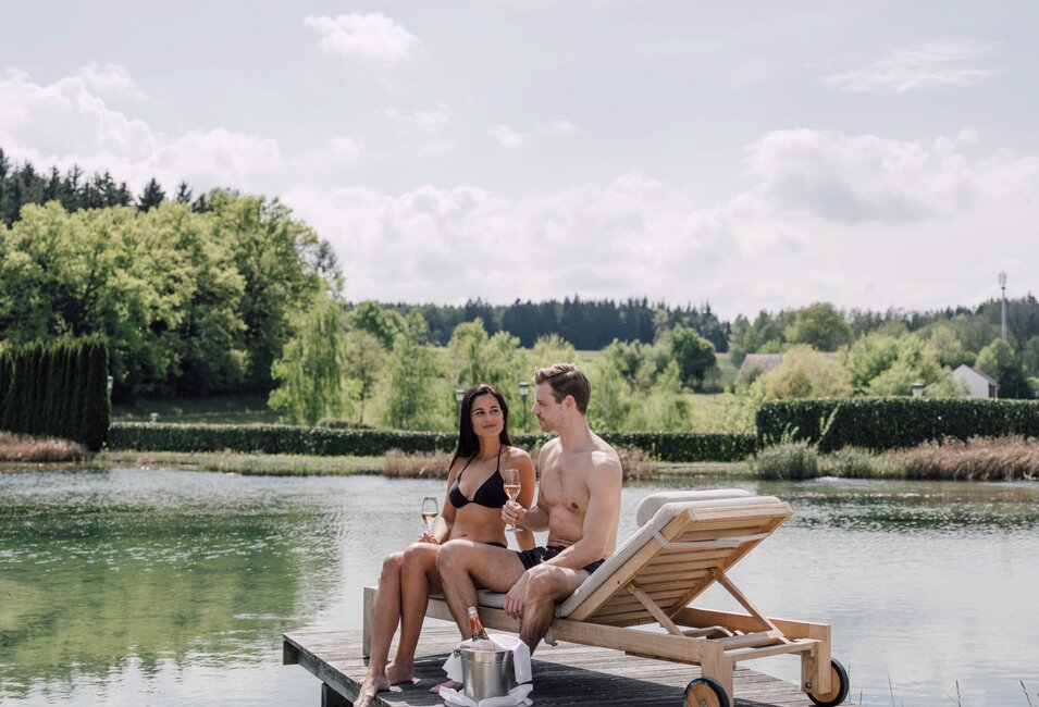 Paar auf Badesteg mit Sekt am See, Liege und grüne Landschaft | © Spa Resort Geinberg / Chris Perkles