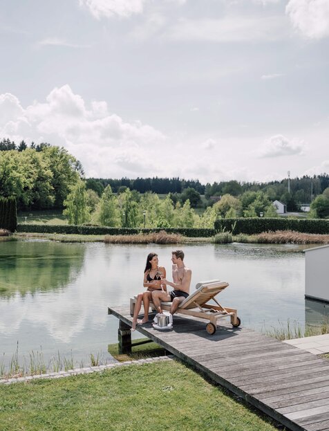 Paar auf Badesteg am See mit Sekt, Liege und grüner Landschaft | © Spa Resort Geinberg / Chris Perkles