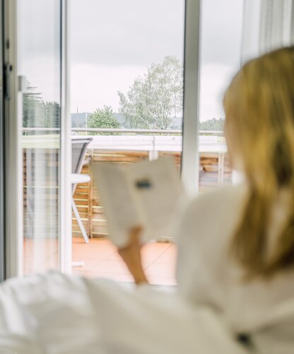 Frau liest ein Buch im Bett des Hotelzimmers mit Blick auf den Balkon | © Spa Resort Geinberg / Chris Perkles