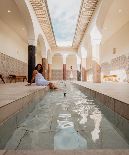 Paar entspannt im offenen Arkadenhof, die Dame lässt die Beine im Wasser baumeln | © Spa Resort Geinberg / Chris Perkles