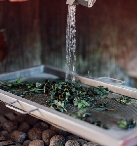 Frische, nasse Kräuterblätter werden auf einem Edelstahlsieb über heißen Saunasteinen mit Wasser beträufelt | © Spa Resort Geinberg / Chris Perkles