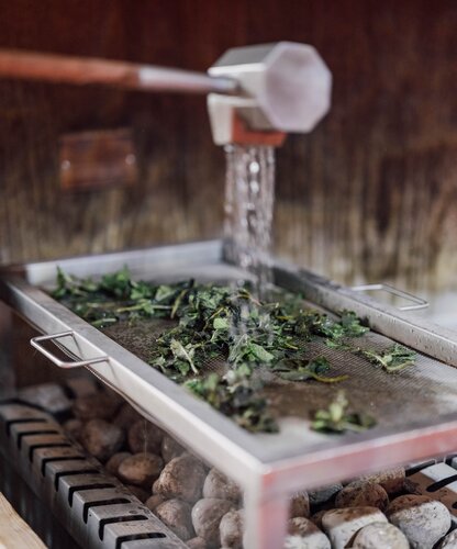 Frische, nasse Kräuterblätter werden auf einem Edelstahlsieb über heißen Saunasteinen mit Wasser beträufelt | © Spa Resort Geinberg / Chris Perkles