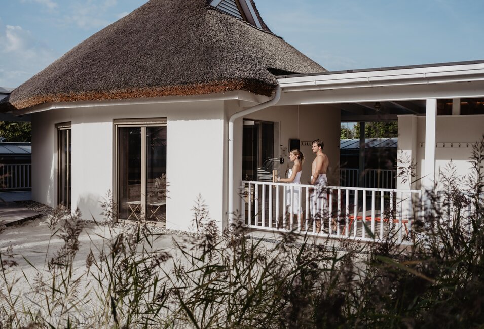Paar in Bademänteln auf Veranda eines Hauses mit Reetdach, im Vordergrund Schilfgras