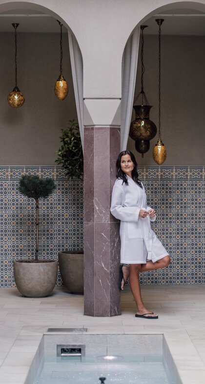 Entspannte Frau lehnt sich an eine Säule im Bademantel im SPAplus Arkadenhof | © Spa Resort Geinberg / Chris Perkles