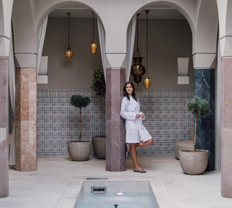 Entspannte Frau lehnt sich an eine Säule im Bademantel im SPAplus Arkadenhof | © Spa Resort Geinberg / Chris Perkles