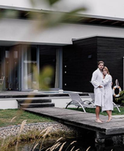 Paar in weißen Bademänteln auf Holzsteg vor einer Villa