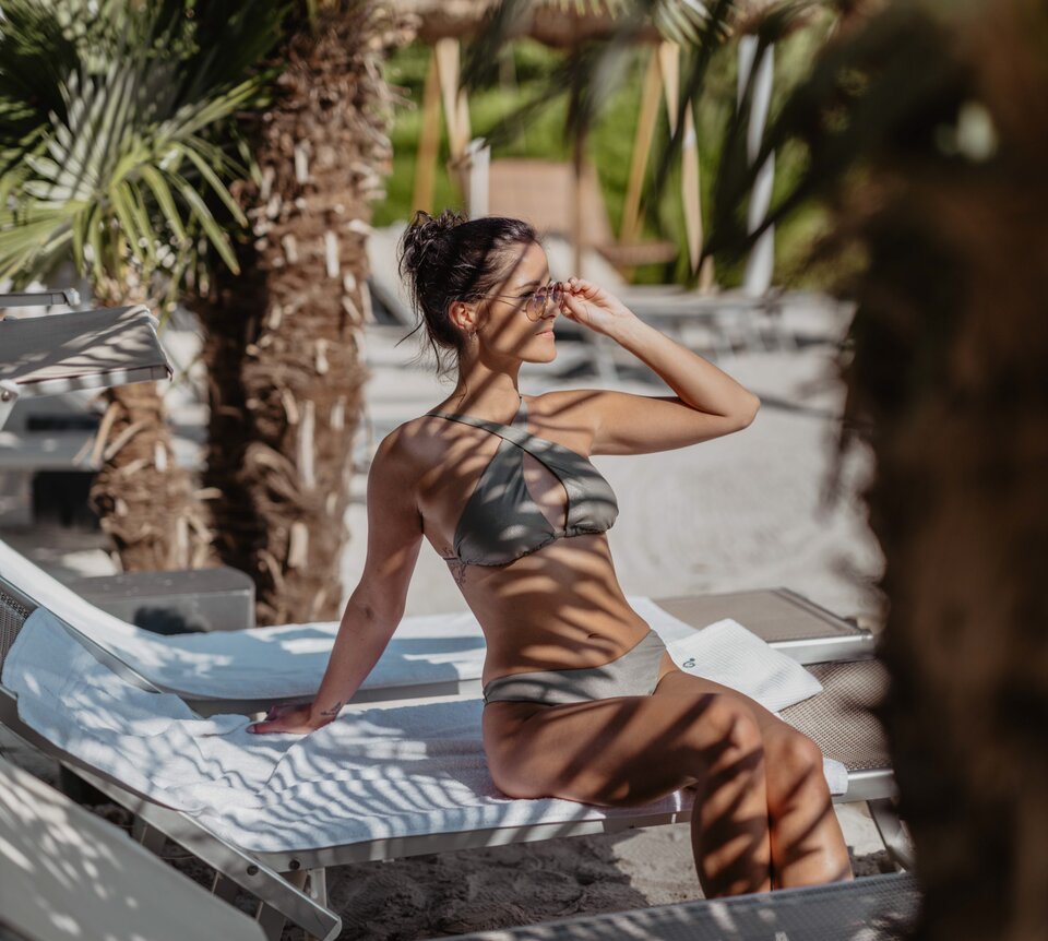Frau im Bikini entspannt auf Liege am Strand unter Palmen in der Karibik Lagune | © Spa Resort Geinberg / Chris Perkles