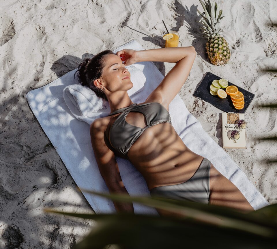 Frau im Bikini liegt am Strand neben Ananas, Cocktail und Obst in der Karibik Lagune | © Spa Resort Geinberg / Chris Perkles