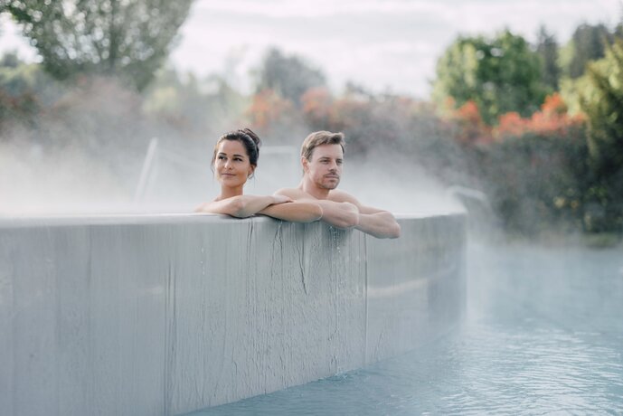 Paar entspannt im Kaskaden-Becken, Dampf steigt auf und im Hintergrund sind farbige Herbstbäume | © Spa Resort Geinberg / Chris Perkles