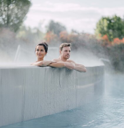 Paar entspannt im Kaskaden-Becken, Dampf steigt auf und im Hintergrund sind farbige Herbstbäume | © Spa Resort Geinberg / Chris Perkles