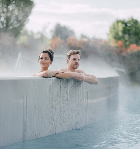 Paar entspannt im Kaskaden-Becken, Dampf steigt auf und im Hintergrund sind farbige Herbstbäume | © Spa Resort Geinberg / Chris Perkles