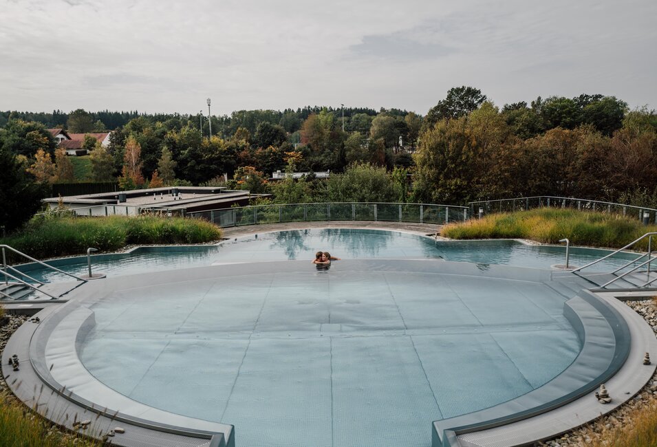 Paar schwimmt im Kaskadenbecken des Spa Resort Geinberg im Herbst vor bewaldeter Landschaft