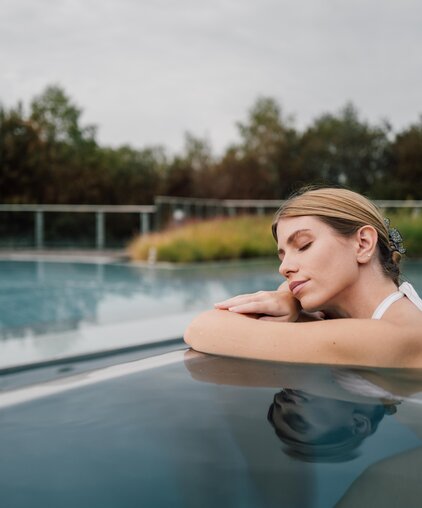 Frau im weißen Bikini entspannt mit geschlossenen Augen im Kaskaden-Becken des Spa Resort Geinberg | © Spa Resort Geinberg / Chris Perkles