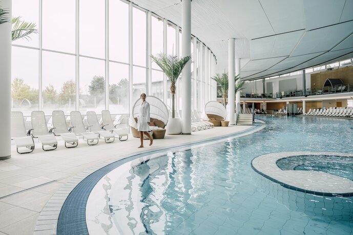 Frau im Bademantel schlendert am Rand des Thermal-Becken indoors in der weitläufigen Therme Geinberg | © Spa Resort Geinberg / Chris Perkles