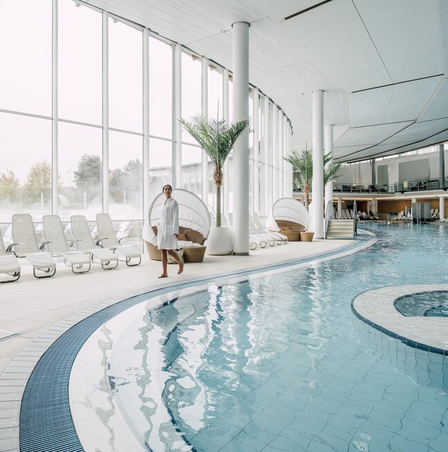 Frau im Bademantel schlendert am Rand des Thermal-Becken indoors in der weitläufigen Therme Geinberg | © Spa Resort Geinberg / Chris Perkles