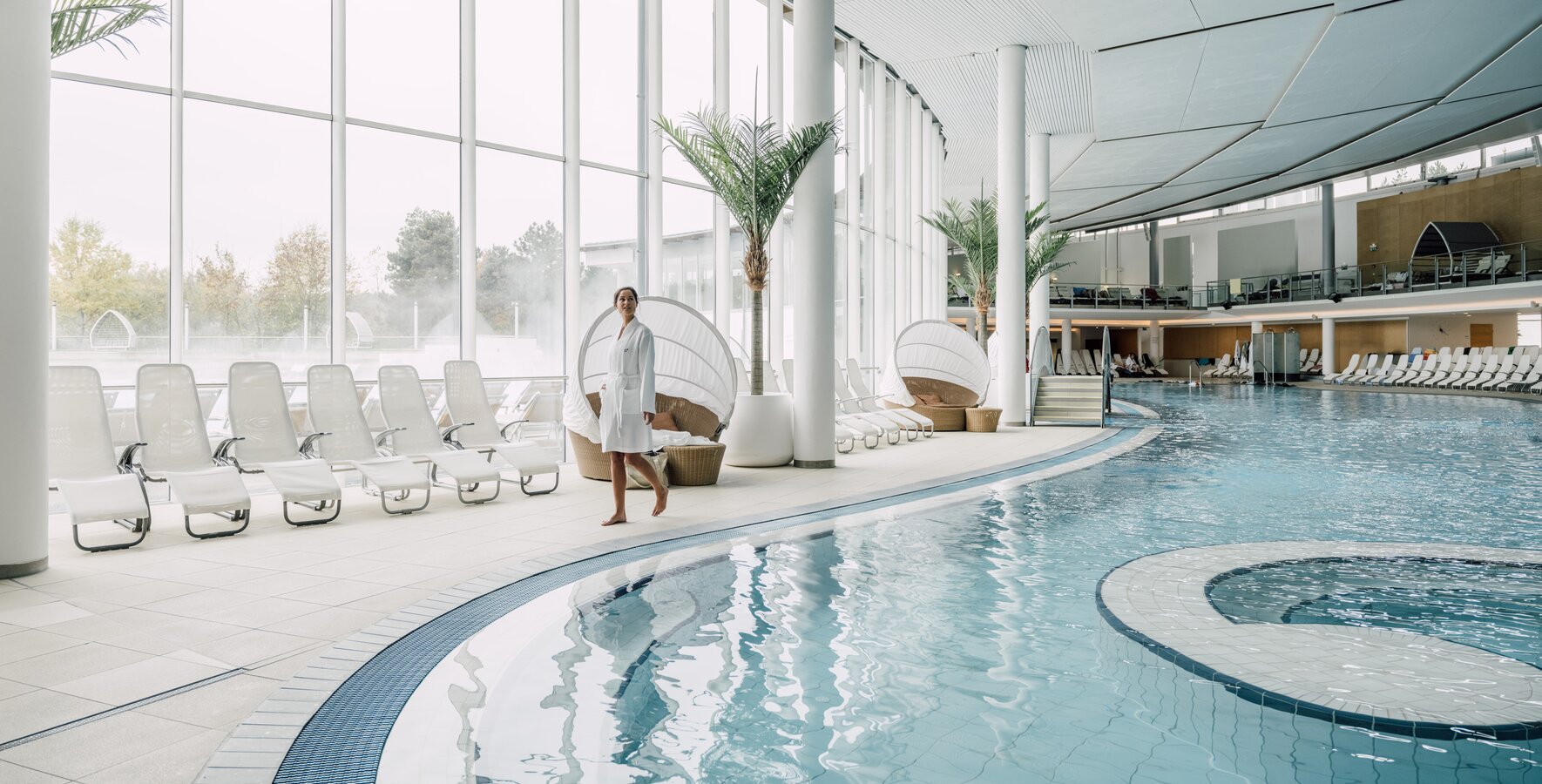 Frau im Bademantel schlendert am Rand des Thermal-Becken indoors in der weitläufigen Therme Geinberg | © Spa Resort Geinberg / Chris Perkles