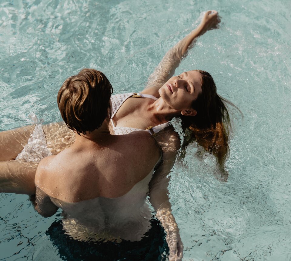 Mann trägt blonde Frau auf Händen im Wasser des Thermal-Beckens outdoor im Spa Resort Geinberg | © Spa Resort Geinberg / Chris Perkles