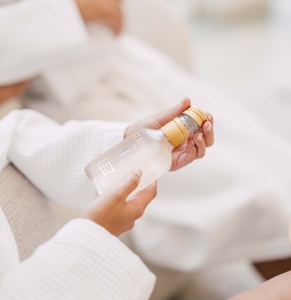 Hände halten eine durchsichtige Flasche mit goldenem Verschluss vor weißem Hintergrund | © Spa Resort Geinberg / Chris Perkles